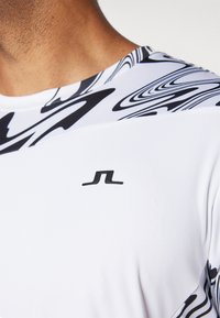 Hvid sportstrøje med et sort abstrakt mønster på skuldrene og et lille sort logo på brystet. Glat stoftekstur.