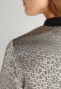 Primo piano della spalla e del collo di una persona che indossa una giacca con trama caratterizzata da motivi circolari metallici argentati e neri, con un colletto nero.