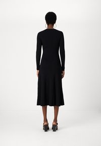 Fekete bordás, hosszú ujjú ruha, melynek testhez simuló felső része van és harangszoknyája midi hosszúságú. Vastag sarkú szandállal és nagy karika fülbevalóval viseli.