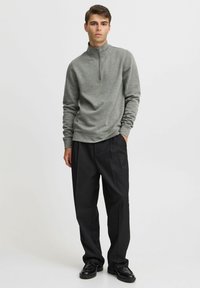Grå zip-up sweatshirt med hög krage, kombinerad med svarta plisserade byxor och svarta skor, framför en enfärgad bakgrund.