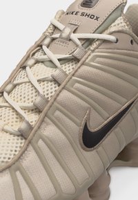Μπεζ αθλητικό παπούτσι Nike Shox με επάνω μέρος από δίχτυ, χαρακτηριστικό κορδόνι, λαστιχένιες ρίγες και μαύρο λογότυπο Nike Swoosh στο πλάι.