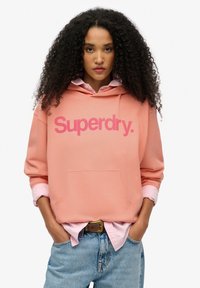 Roze hoodie met "Superdry." logo, kangoeroezak en oversized pasvorm, gelaagd over een lichtroze shirt en gecombineerd met blauwe jeans.