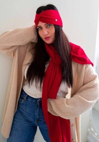 Un bandeau rouge et une écharpe en tissu doux, portés avec un cardigan en maille beige, un top blanc et un jean bleu. Présente un design torsadé et un accent de bouton.