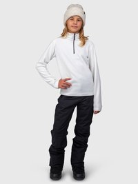 Ragazza in piedi con una mano sui fianchi, che indossa un fleece bianco con zip, pantaloni neri, stivali neri e un berretto di maglia beige su uno sfondo semplice.