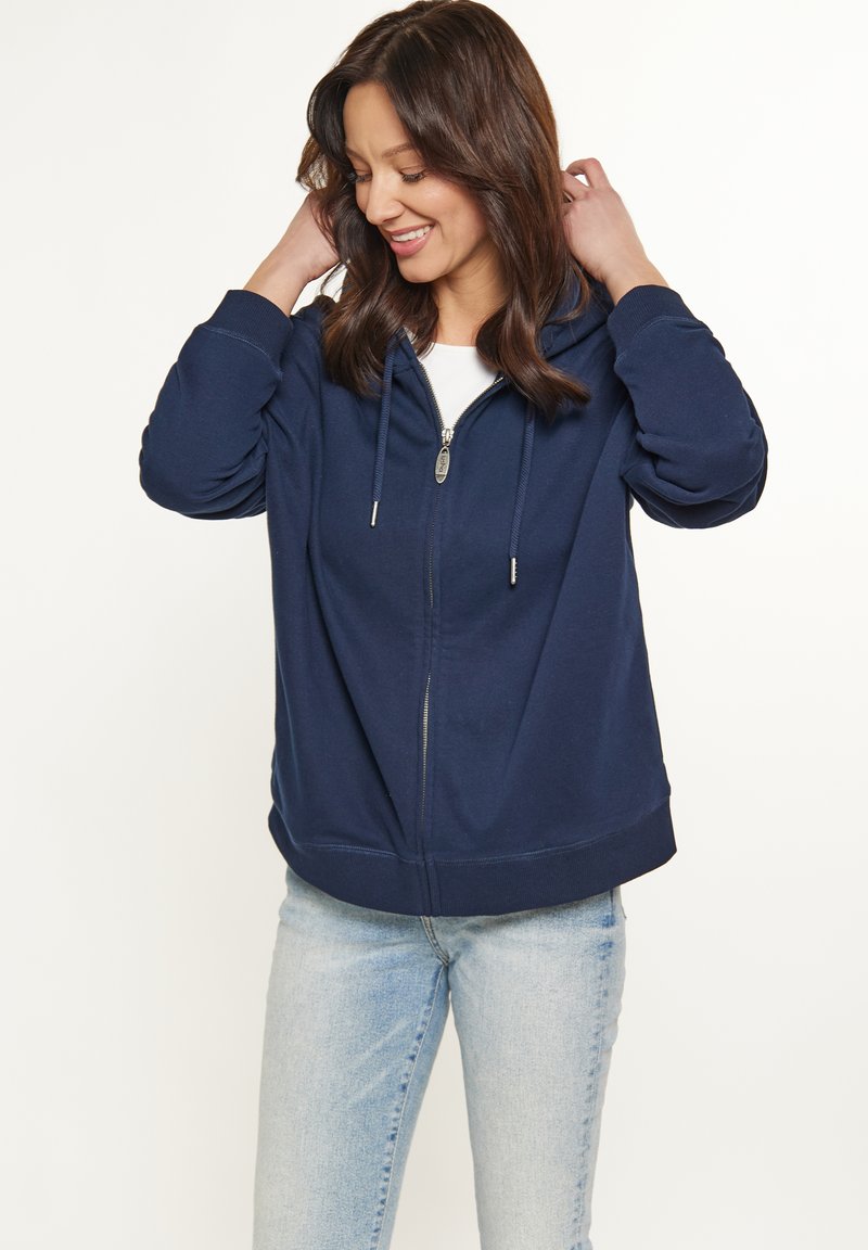 Felpa con zip di colore blu navy in materiale morbido, dotata di cappuccio, tasche laterali e orlo a costine. Abbinata a jeans azzurri chiari.