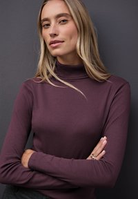 Maglione a collo alto ribbed color bordeaux con maniche lunghe, caratterizzato da un design aderente e una texture morbida. Indossato con le braccia incrociate e un anello d'argento.