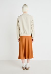 Beige bomberjakke med en glatt tekstur, elastisk midje, sammen med et rustfarget midiskjørt. Hvite sokker og brune sandaler synlige.