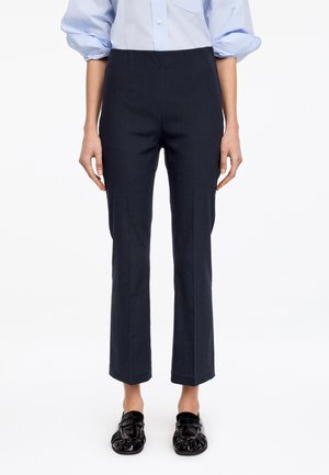 Frau trägt schwarze gerade geschnittene Hose, hellblaues Hemd mit Knöpfen und hochgekrempelten Ärmeln sowie schwarze glänzende Loafers, steht vor weißem Hintergrund.