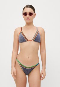 Bikini-Set mit einem dreieckigen Oberteil und passendem Unterteil, versehen mit mehrfarbigem Zickzack-Stich auf einem dunklen glitzernden Stoff. Begleitet von einer rosa Sonnenbrille.
