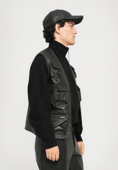 Man in black leather cap, black turtleneck sweater, black leather vest with multiple pockets, and black leather pants, standing in profile.
