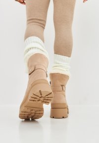 Torby w kolorze tan z zamszu z teksturowaną gumową podeszwą, z paskiem z metalowym guzikiem oraz pluszowym wykończeniem na górze.