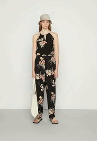 Macacão preto com flores, pernas largas, alças finas e um detalhe em forma de chave na gola. Combinado com um chapéu balde cinza claro e uma mala tote branca.