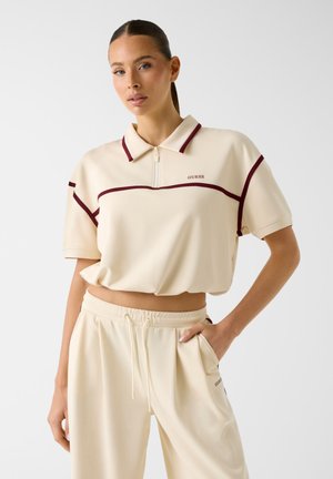 Femme portant un polo court beige avec bordure bordeaux et un pantalon beige assorti à cordon de serrage, main dans la poche.