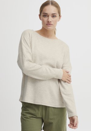 Beige langermet strikket topp med båtutringning og avslappet passform. Teksturert stoff med synlige flekker. Sammen med olivengrønne shorts.
