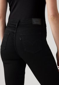 Jeans negros de cintura alta con una textura suave, costuras sutiles, dos bolsillos traseros y una distintiva etiqueta de Levi Strauss en la cinturilla.