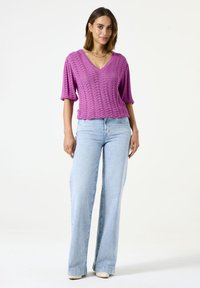 Paarse gebreide top met een V-hals, korte mouwen en een textuurpatroon. Gecombineerd met lichtblauwe wijde jeans en beige schoenen.