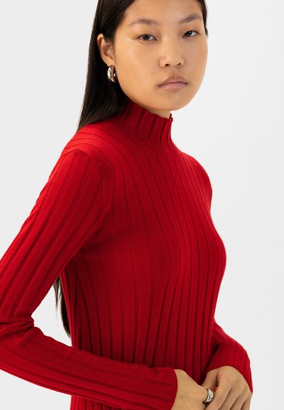 Roter gerippter Pullover mit hohem Kragen und langen Ärmeln, der vertikale Streifen für Textur präsentiert. Aus weichem Strickmaterial und körpernaher Passform.