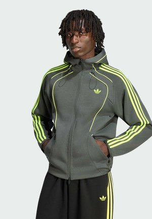 Person trägt einen dunkelgrauen Zip-Hoodie mit neongelben Streifen und Adidas-Logo, Hände in den Taschen und passende schwarze Hose mit gelben Streifen.