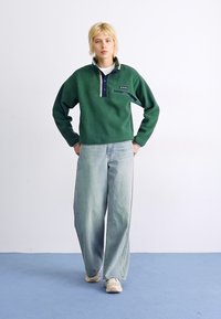 Pullover en polaire vert avec un col montant, accents bleu marine et une poche zippée ; associé à un jean large bleu clair et des baskets blanches.