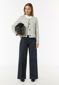 Witte jas met zwarte strepen en knopen, gecombineerd met donkere wijde jeans met contrasterende stitching. Model houdt een zwarte tas vast.