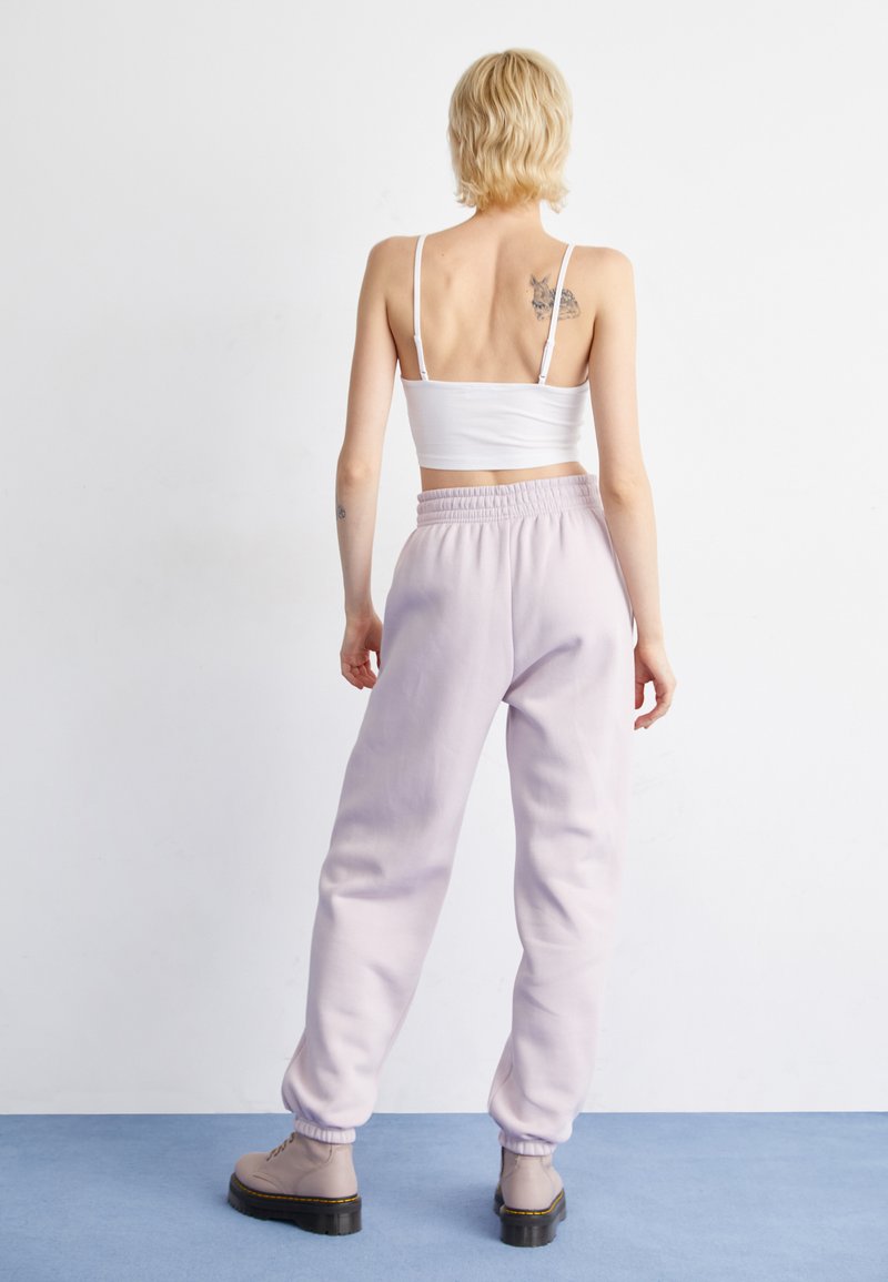 Person, der ein weißes Crop-Top, helllila Jogginghosen und beigen Stiefeln trägt, steht mit dem Rücken an einer weißen Wand und zeigt Rückentätowierungen.
