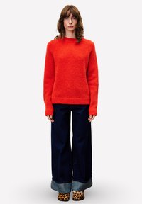 Frau steht in einem leuchtend roten Pullover, weiten, hochgekrempelten dunkelblauen Jeans und Leoprint-Slippern vor einem weißen Hintergrund.