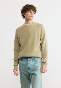 Jeune homme portant un pull beige texturé et un jean bleu clair, debout et regardant sur le côté sur un fond blanc uni.