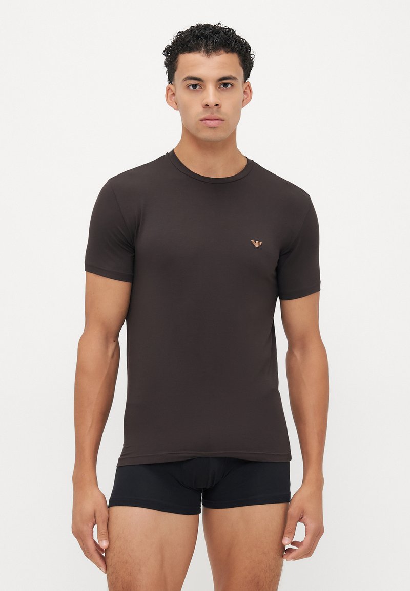 T-shirt ajustada castanha, feita de tecido suave, com decote redondo e mangas curtas; inclui um pequeno logotipo na área do peito esquerdo.