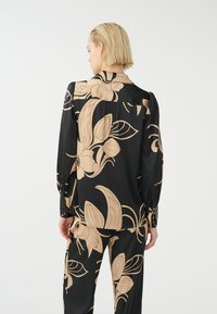 Blusa a maniche lunghe nera con ampio motivo floreale beige, caratterizzata da polsini con bottoni e una vestibilità rilassata. Tessuto liscio con una leggera lucentezza.