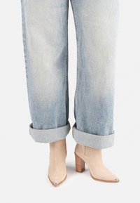 Botas de tobillo de ante beige con puntera afilada y un acento plateado en la punta. Combinadas con jeans de mezclilla de pierna ancha y puños en azul claro.