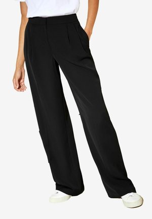 Personne portant un pantalon noir ample et des baskets blanches, debout avec une main partiellement dans la poche, sur un fond uni.
