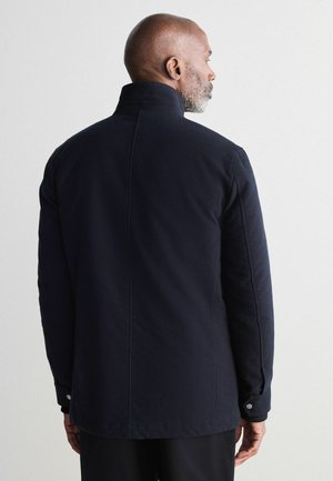 Chaqueta azul marino con un cuello alto, diseño sencillo, textura de la tela y detalles de botones a presión en las mangas. Vista trasera mostrada.