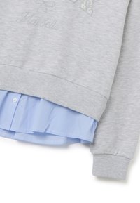 Sudadera gris con puños de canalé, con bordado de "Ma Belle"; superpuesta con el dobladillo de una camisa a rayas azul claro.