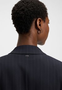 Navy pinstripe blazer with a notched collar. Features a subtle metallic accent at the back and visible seam details. Textured fabric.