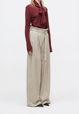 Femme portant un chemisier manches longues rouge foncé avec un nœud et un pantalon beige large taille haute avec ceinture, debout devant un fond uni.