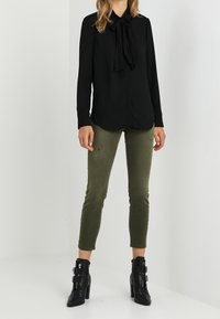 Blusa negra con lazo, jeans slim fit verde oliva y botas negras de tobillo con detalles de hebilla. Tela lisa, diseño ajustado.