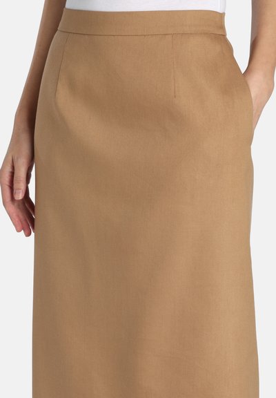 Kvinde iført en knælængde nederdel i kamelfarve med sidelommer, kombineret med en hvid top, stående foran en hvid baggrund.