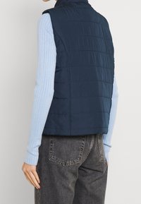 Navy quilted vest with a high collar and no sleeves, paired with a light blue ribbed long-sleeve shirt and dark denim jeans.