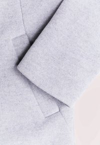 Světle šedá vlna na kabát s jemnou texturou, vybavená přeloženým rukávem a čistým, minimalistickým designem. Bez viditelných kovových prvků či zdobení.