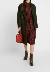 Abrigo de lana verde oliva, vestido de diseño burdeos, bolso rojo y botines de piel de cocodrilo verde oscuro y brillante.