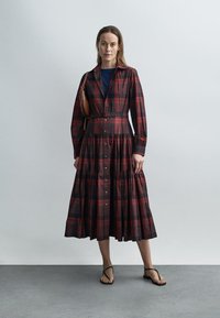 Rochie cu nasturi, mâneci lungi, în carouri roșu închis și negru, cu fustă în straturi și talie cu curea. Purta cu papuci flip-flop negri.