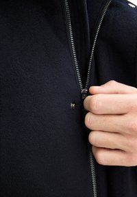 Navy villatakki, jossa on näkyvä hopeinen vetoketju ja nappilukko. Kangas on pehmeä ja tekstuuri tuo esiin istuvan muotoilun.