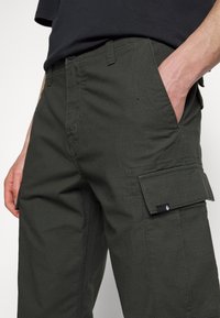 Man i mörkgröna cargoshorts med händerna i framfickorna och en svart skjorta delvis synlig.