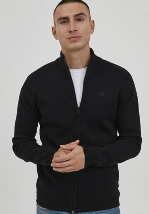 Black zip-up cardigan made of knit fabric, featuring a mock neck, ribbed cuffs, and a subtle logo detail on the chest.