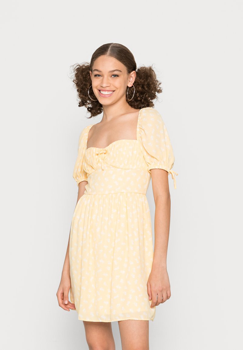 Vestido amarelo floral com mangas curtas bufantes, decote quadrado e detalhe com cordão. Tecido leve, saia rodada e padrão de flores brancas.