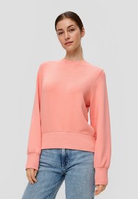 Jonge vrouw poseert in een koraalroze sweatshirt en lichtblauwe jeans tegen een effen lichtgrijze achtergrond.