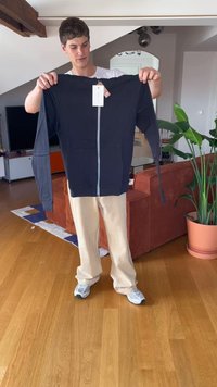 Man som står i vardagsrummet och håller upp en marinblå zip-up hoodie med prislappar fastsatta, beige byxor, vita sneakers och inomhusväxter i närheten.