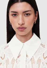 Balta pogām aiztaisāma krekla ar teksturētu mežģīņu dizainu, tai ir ass apkaklis un visā kreklā ir ziedu raksti. Modelis valkā zelta riņķu auskarus.