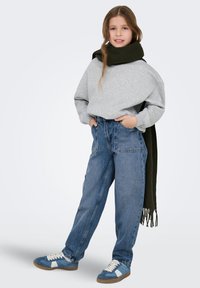 Szürke túlméretezett pulóver, kék magasderekú farmernadrág és kék, fehér részletekkel ellátott tornacipők. Sötétzöld sál a vállakon átvetve.