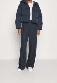 Doudoune bleu marine avec capuche et motif matelassé, associée à un pantalon bleu marine ample avec des bandes blanches sur les côtés. Des baskets blanches complètent le look.